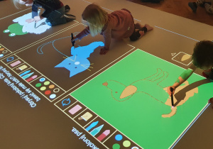Troje dzieci bawi się na podłodze interaktywnej. Dzieci rysują po śladzie oraz kolorują wybrane przez siebie obrazki.
